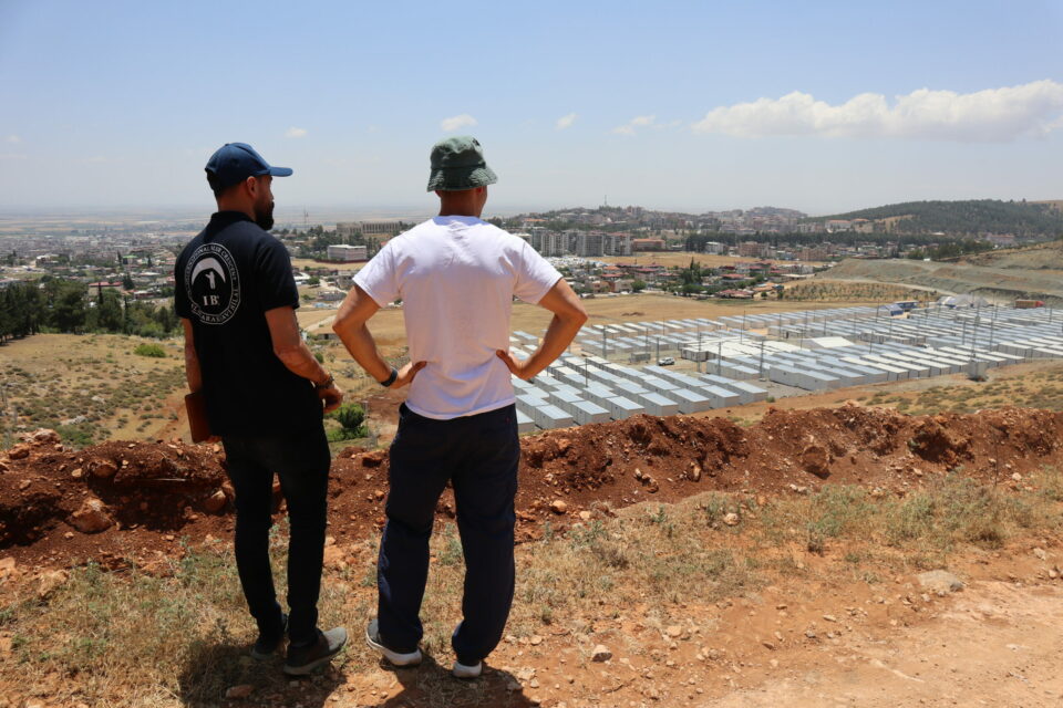 Two men on top of a hill overlooking a turkish city, with hundreds of homes made from shipping containers in the foreground